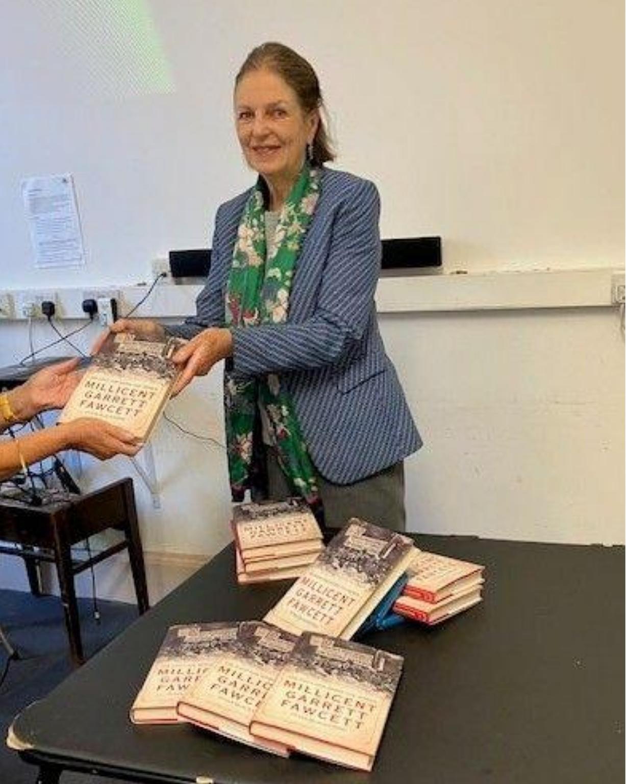 Baroness Tessa Blackstone smiling as she signs copies of her book on Millicent Garrett Fawcett, with more copies stacked on a table